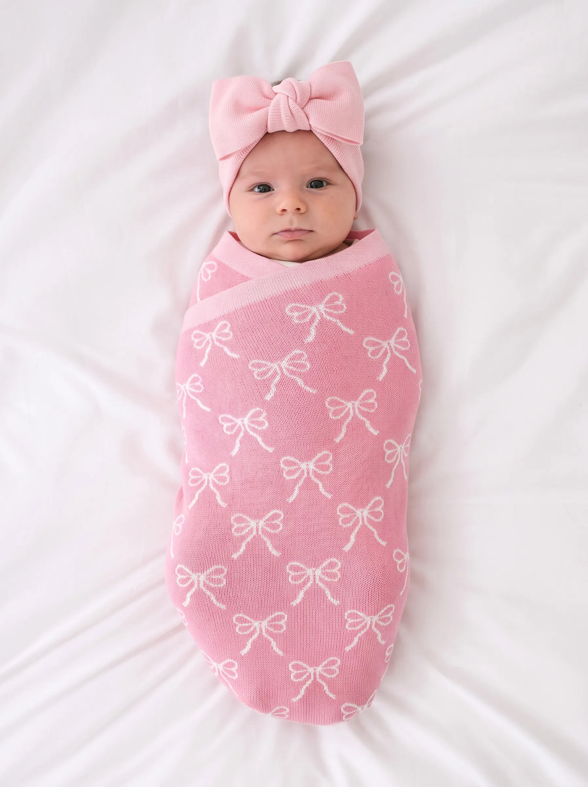 Pink knitted baby swaddle blanket with white bow pattern and matching bow hat.