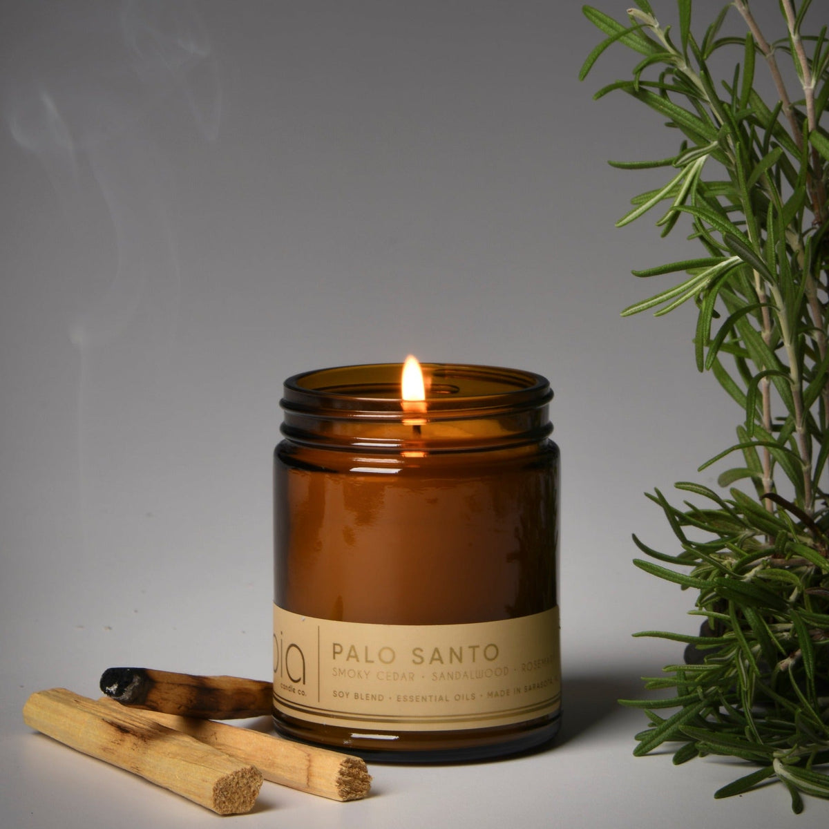 Amber glass soy candle with flame next to wood sticks and green rosemary sprig