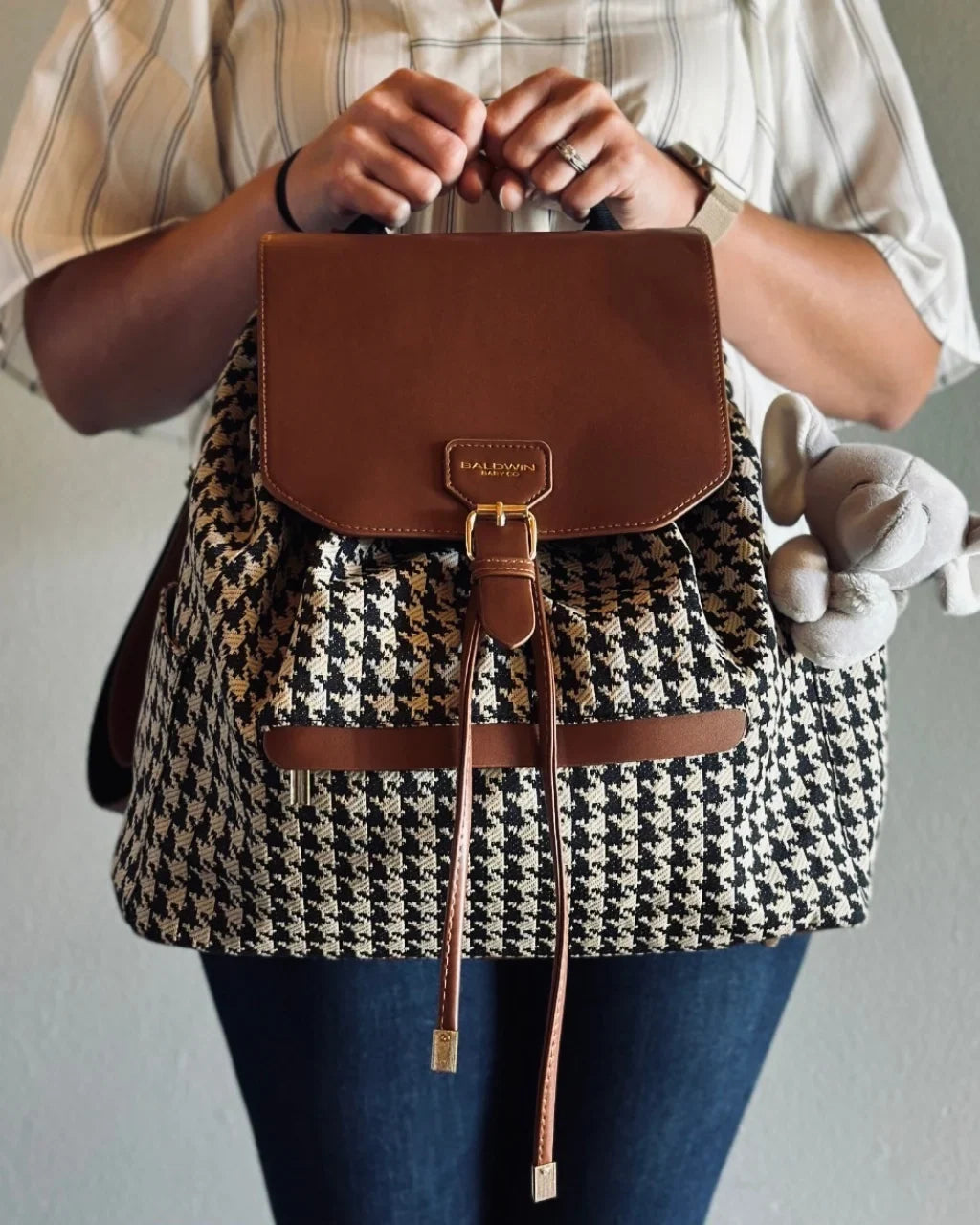 Houndstooth diaper bag with brown leather-look flap and drawstring, carried by its top handle