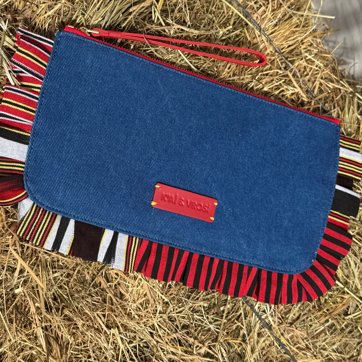 Denim clutch bag with blue flap, red wrist strap, striped ruffle trim, leather logo patch