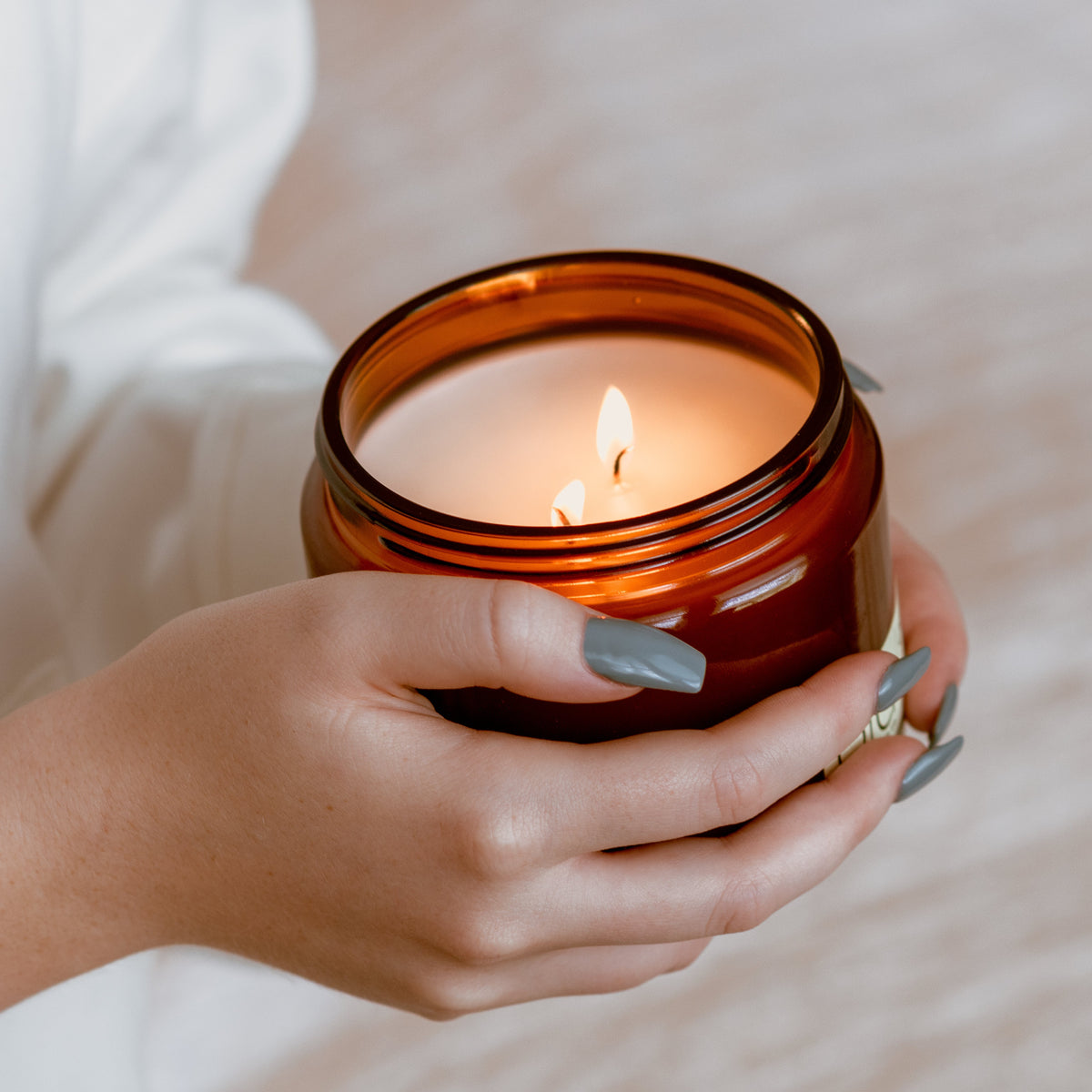 Lit amber soy candle in glass jar held in hands, flame visible through jar