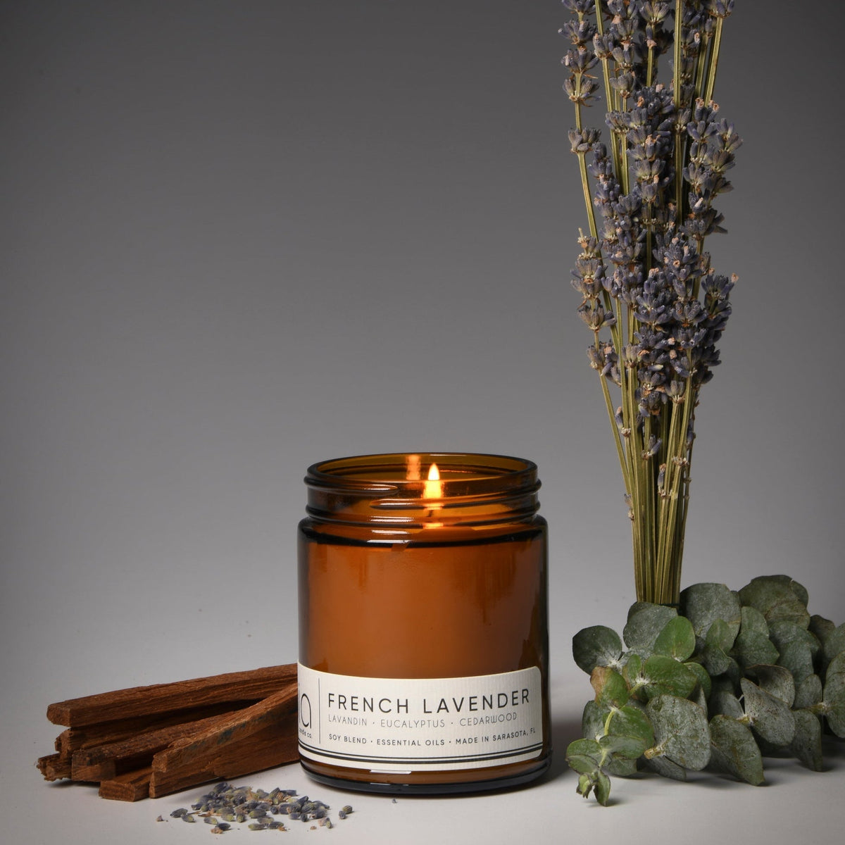 Amber soy candle jar with lit flame beside lavender stems, eucalyptus leaves, and cedar pieces
