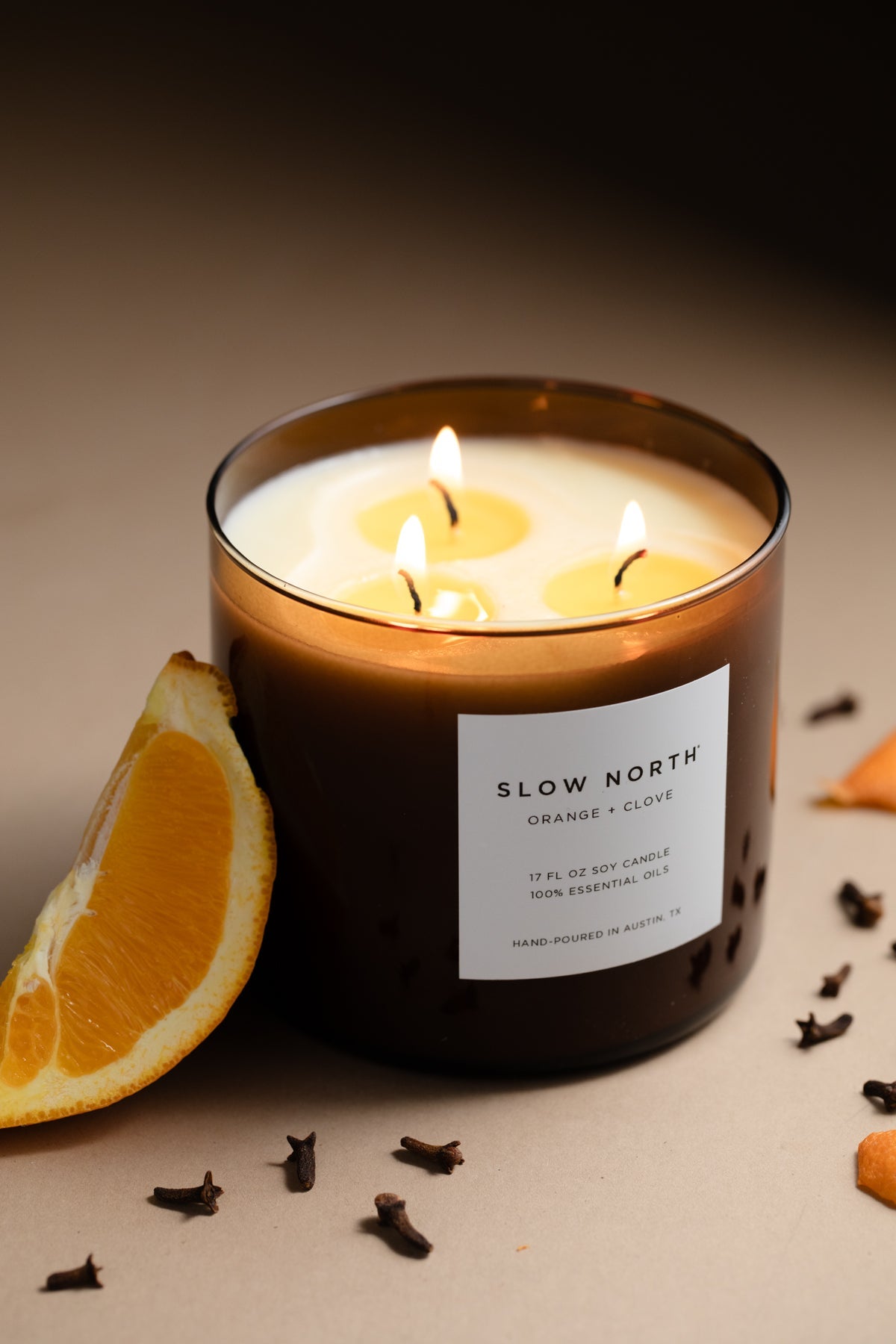 Slow North 3-wick candle in amber glass with orange-clove label and three flames