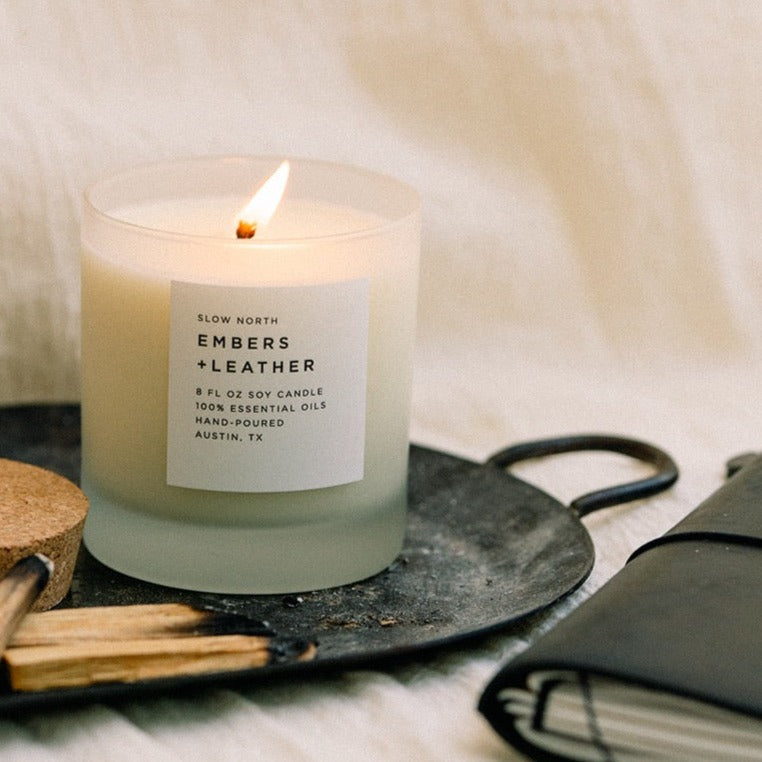 Lit Embers + Leather candle in a pale frosted glass jar with a white label.