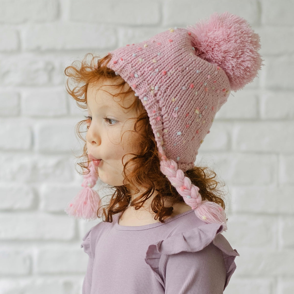 Pink flecked knit earflap hat with pom-pom and braided ties, worn in profile