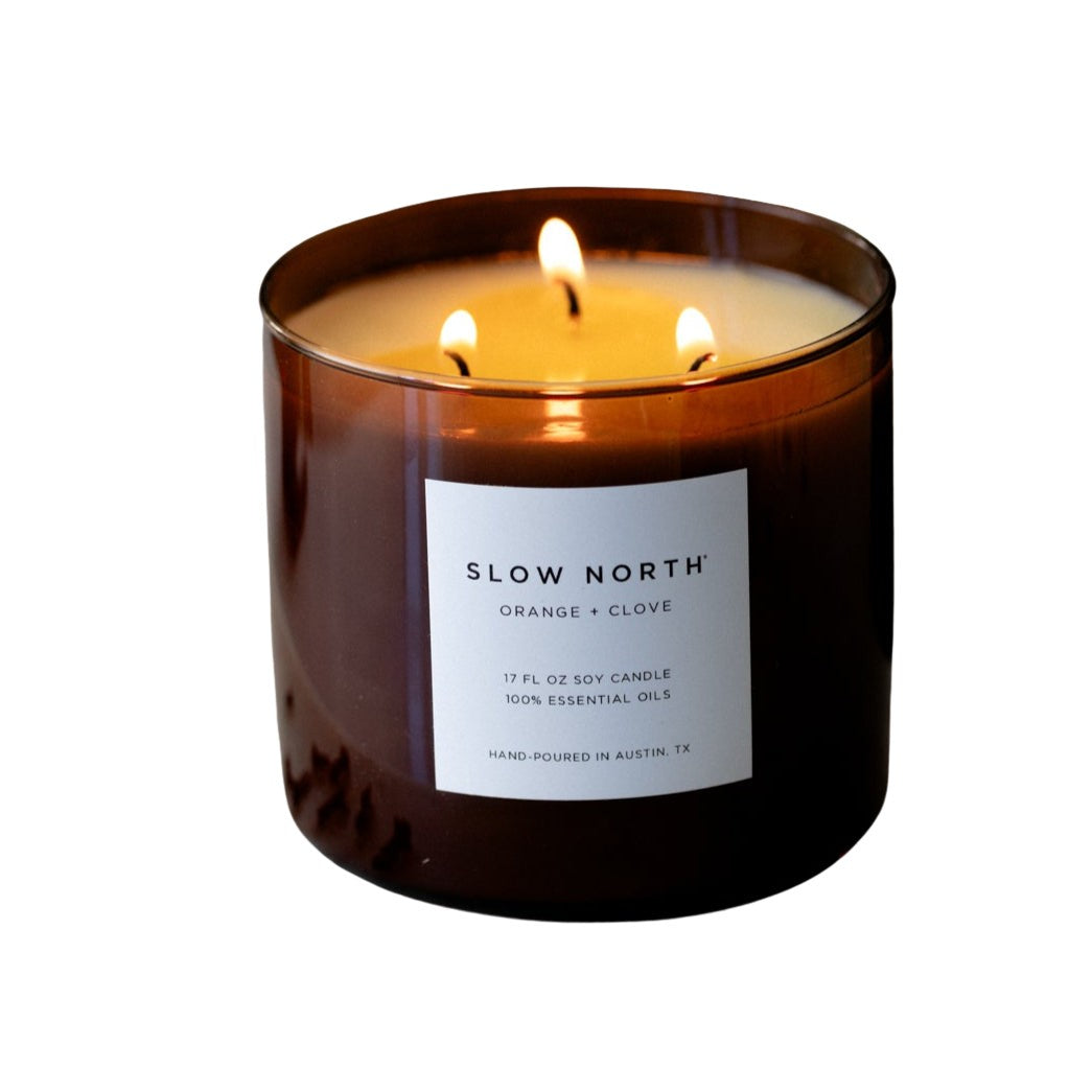 Slow North 3-wick candle in amber glass with orange-clove label and three flames