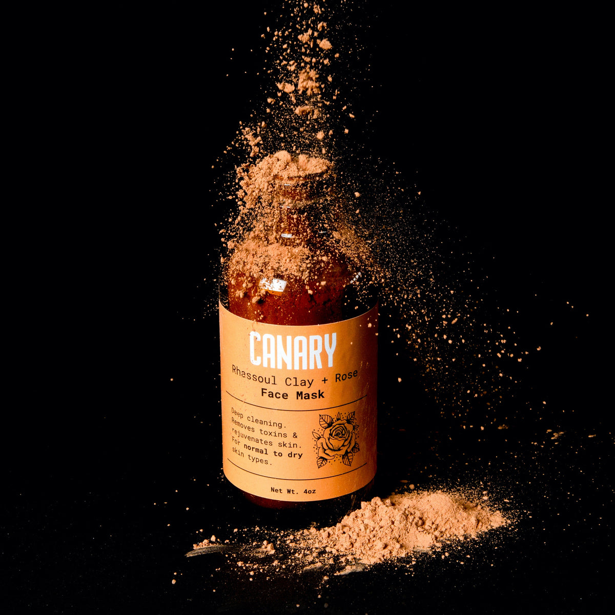 Canary Rhassoul clay and rose face mask bottle beside a mound of clay powder