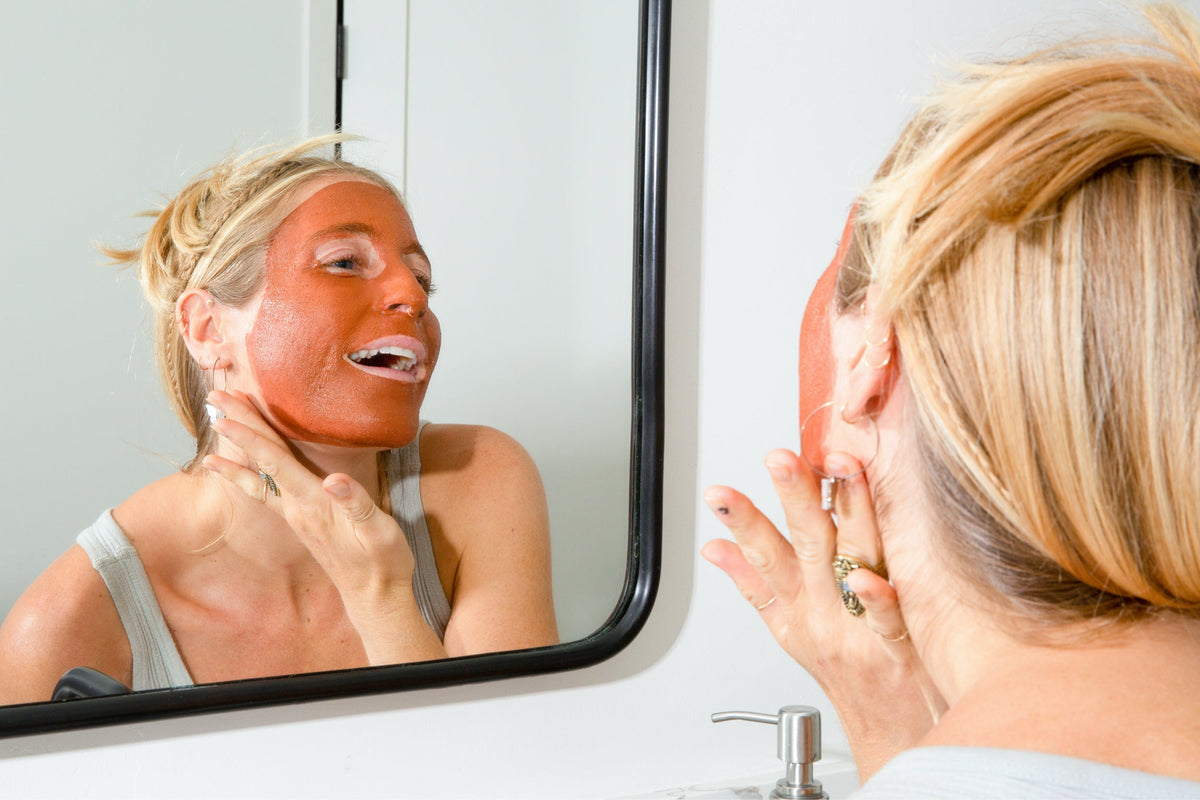 Woman wearing an orange clay face mask and touching her cheek