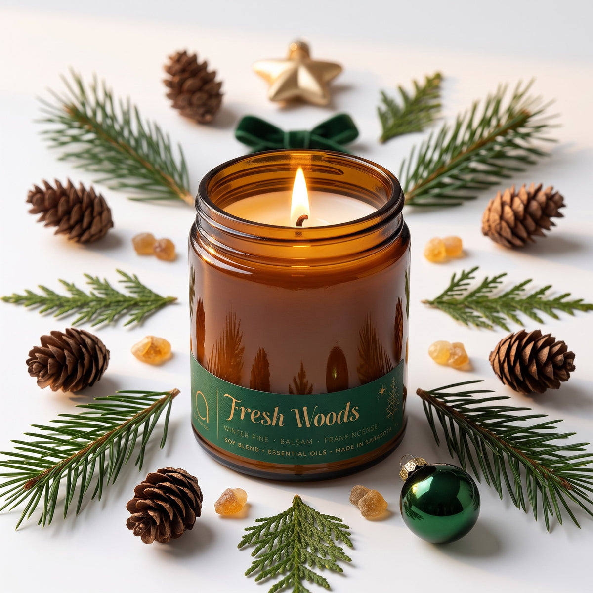 Amber glass candle jar with lit wick and green Fresh Woods label, on greenery