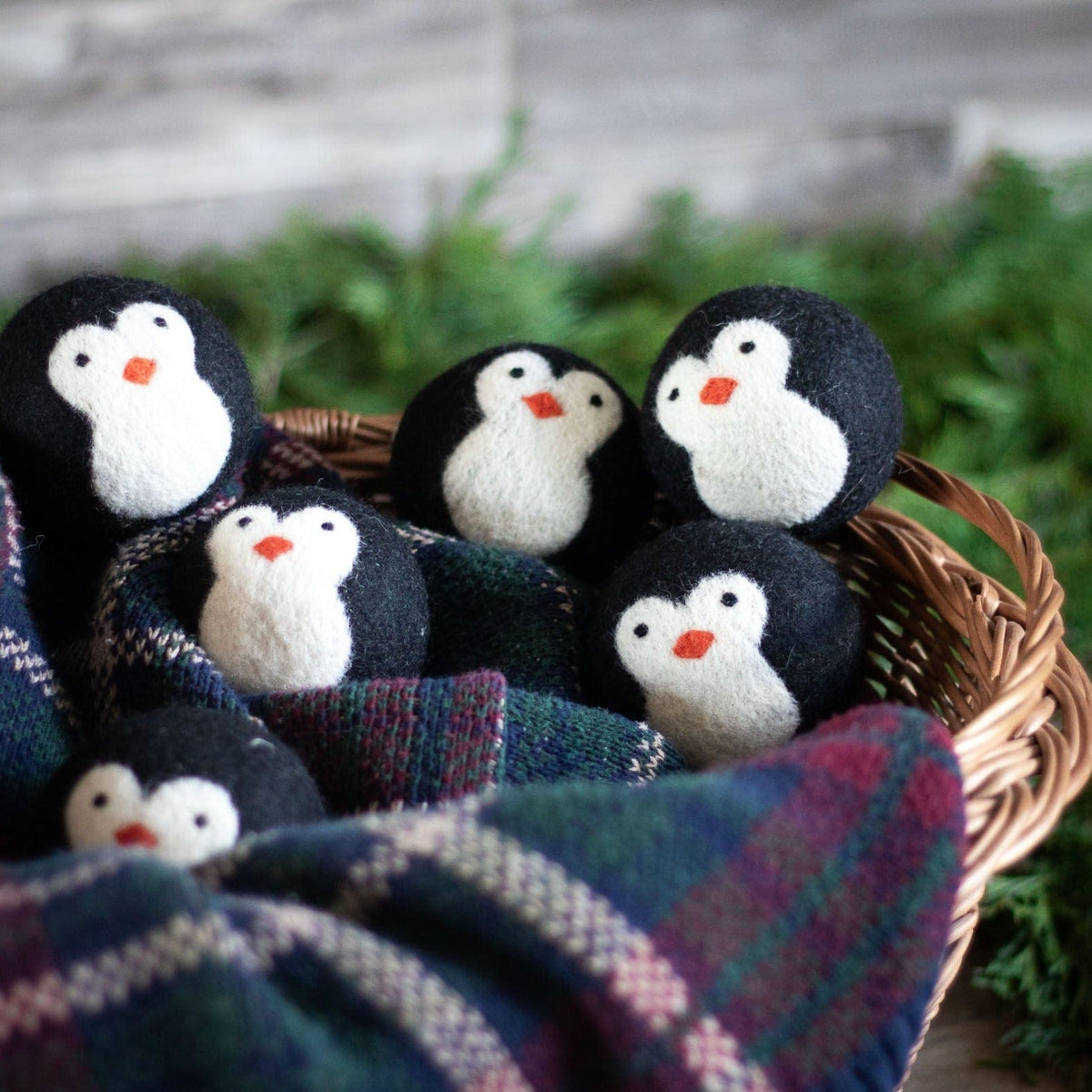 Penguin dryer balls arranged in a wicker basket with a plaid cloth and greenery