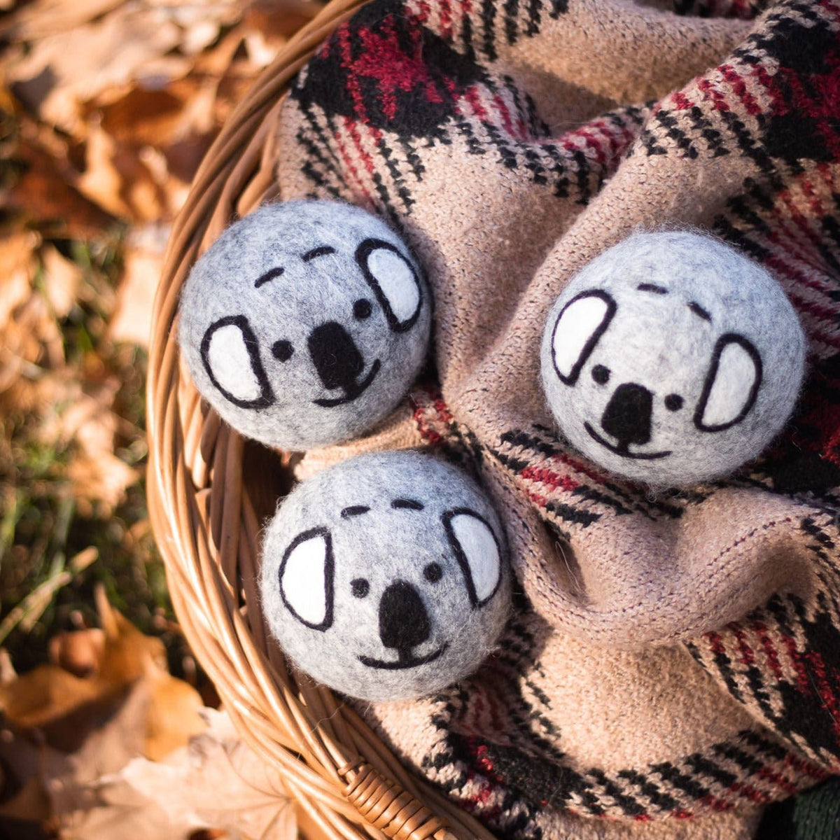 Three gray koala plush balls on a plaid blanket in a wicker basket outdoors