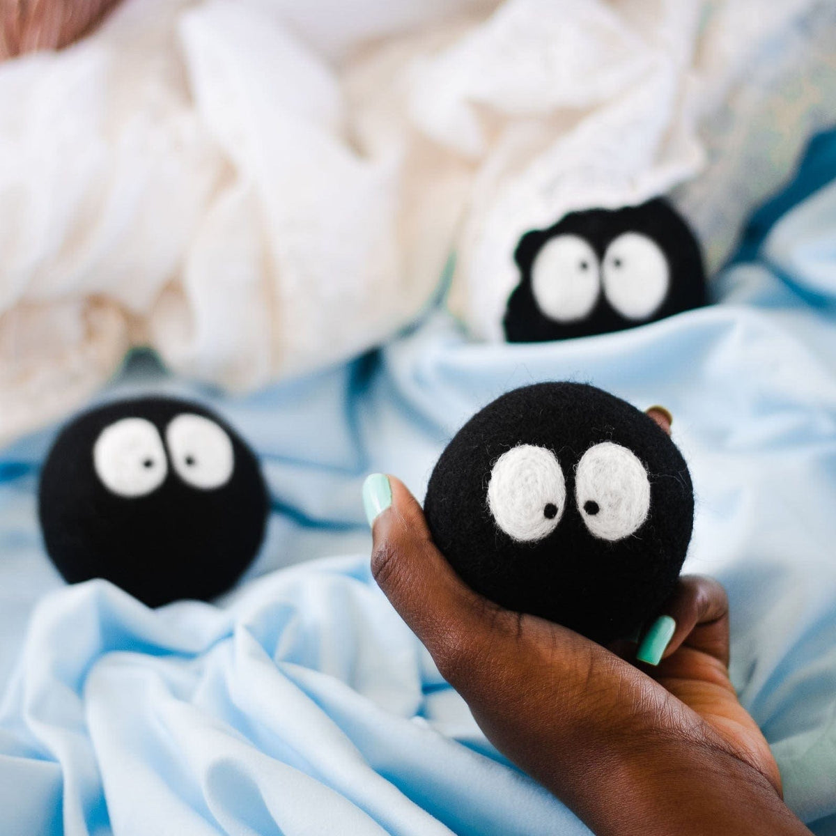 Hand holding a plush black sheep-faced laundry dryer ball on light blue bedding