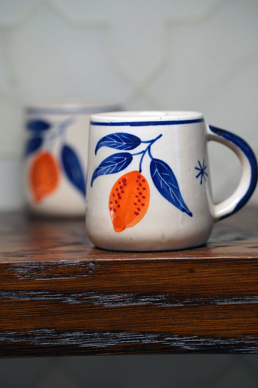 Ceramic coffee mug with white base and blue rim, featuring orange lemon illustration