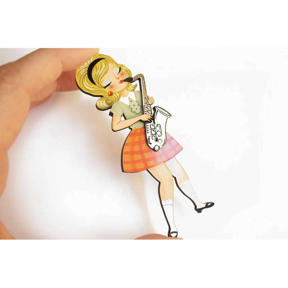 Girl playing saxophone brooch held by hand, yellow hair, green top, orange plaid skirt.