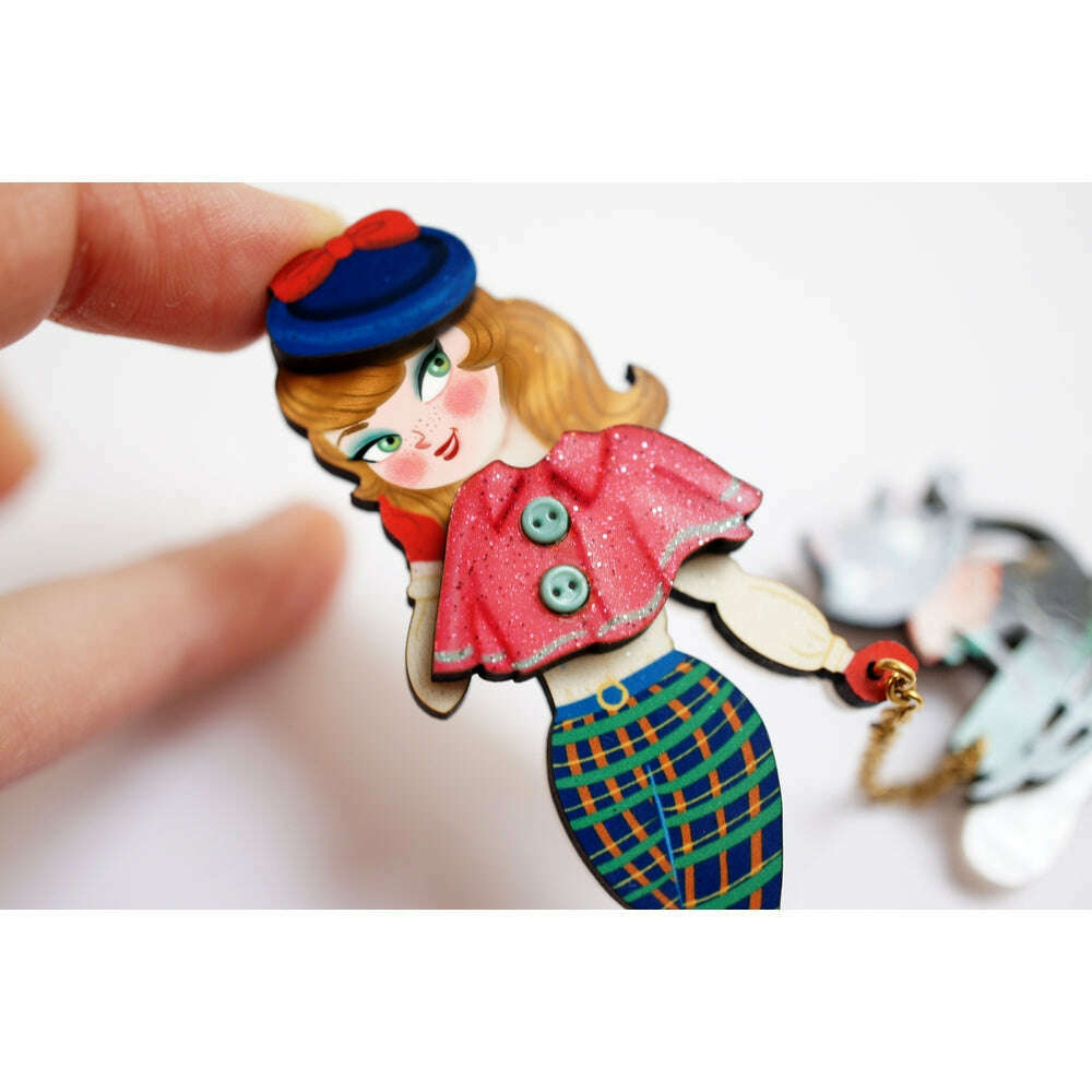 Hand holding enamel brooch: girl with blue hat, cat on sled