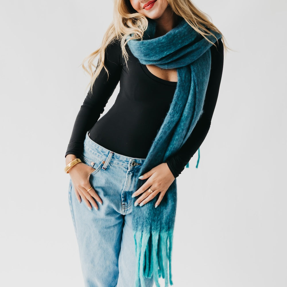 Teal blanket scarf with fringe, worn around the neck over a black top