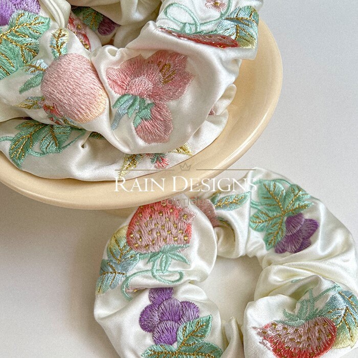 Close-up fruit embroidery scrunchie: strawberries and grapes on ivory ruffled fabric.