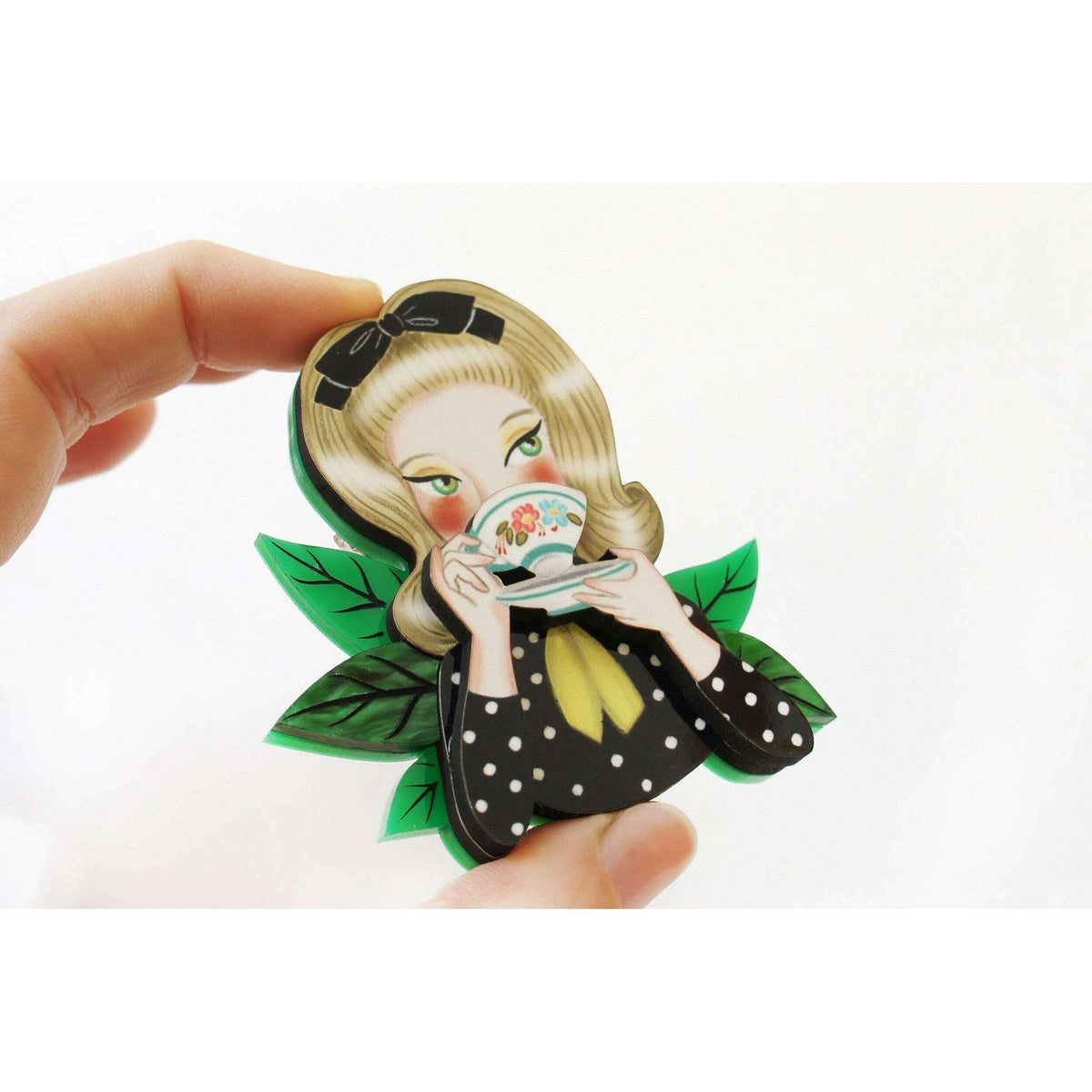 Girl tea-drinking brooch held close-up, showing face, cup, and green leaves