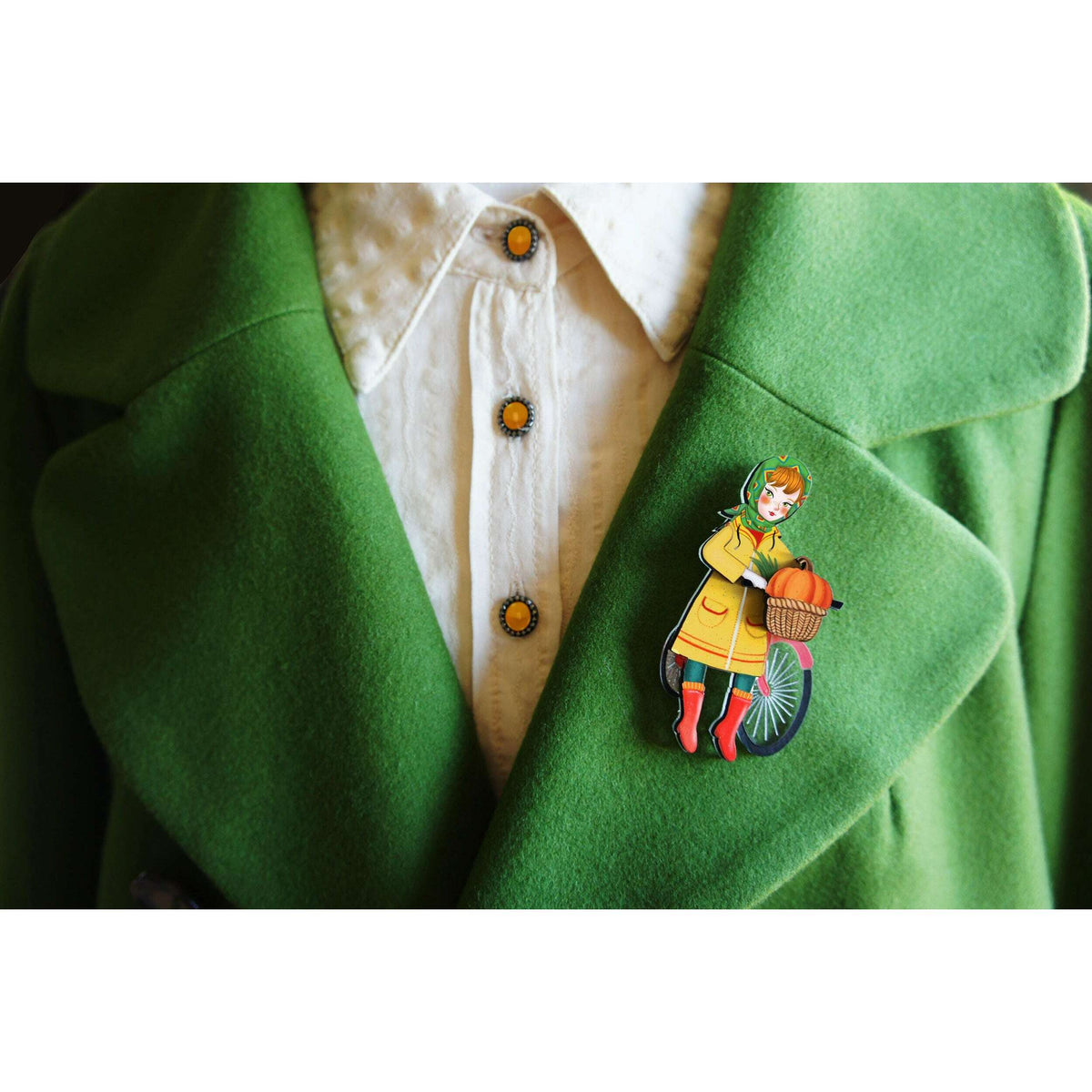 Bicycle girl brooch pinned to green coat lapel, with orange pumpkin basket and bike wheel