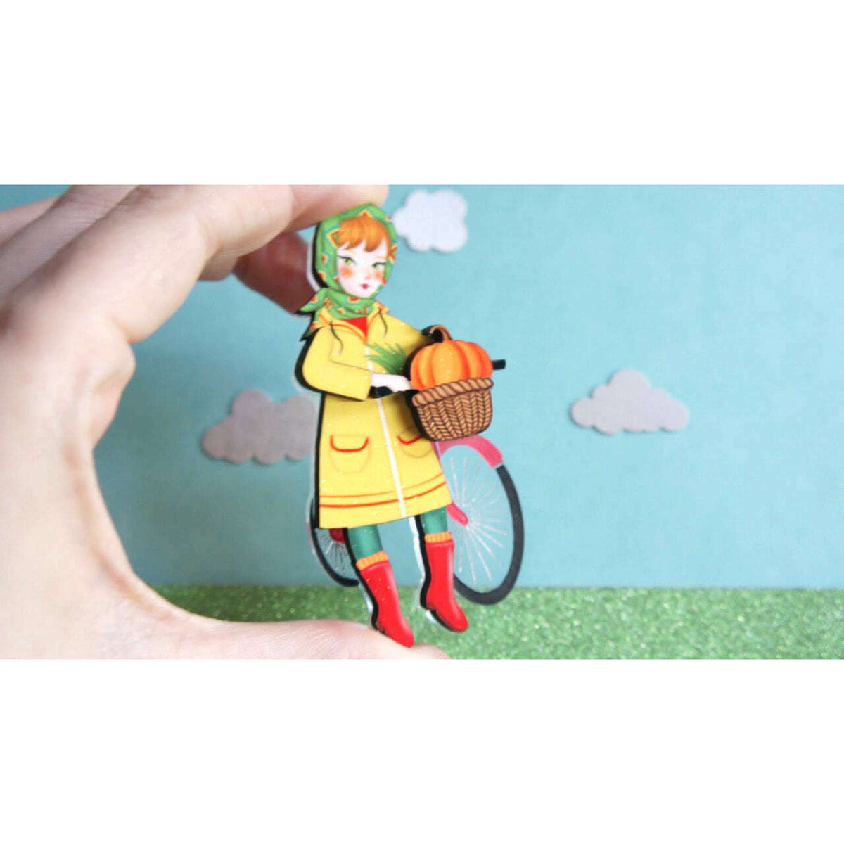 Bicycle girl brooch held between fingers, hooded child with orange pumpkin basket