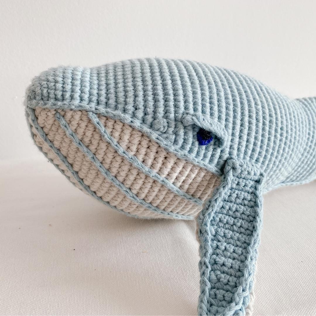 Close-up of a misty blue crocheted whale plush with a cream striped belly and blue eye.