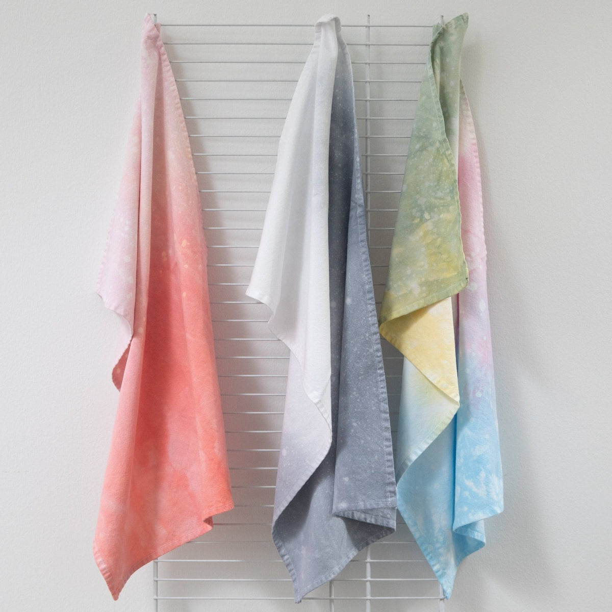 Hand-dyed tea towels hanging on a rack in pastel pink, white, gray, green, blue, and yellow.