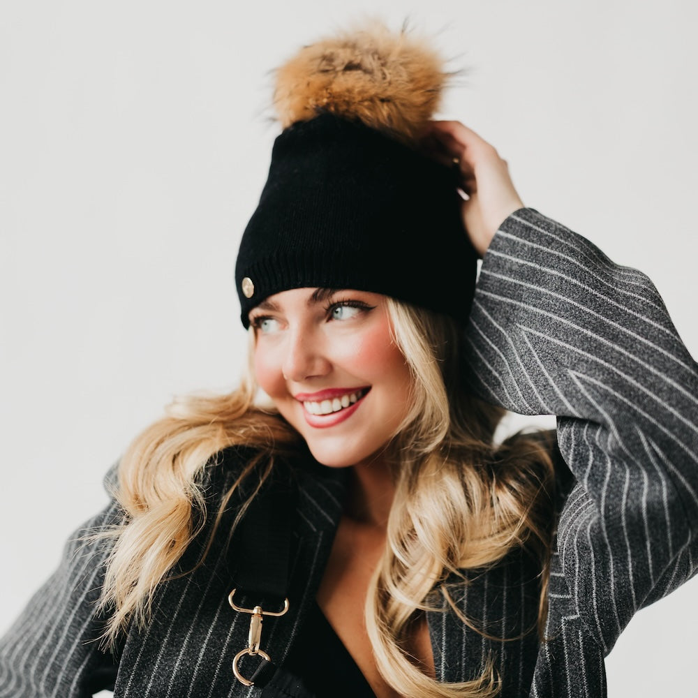 Black knit slouchy beanie with ribbed cuff, gold-tone button, and tan fur pom