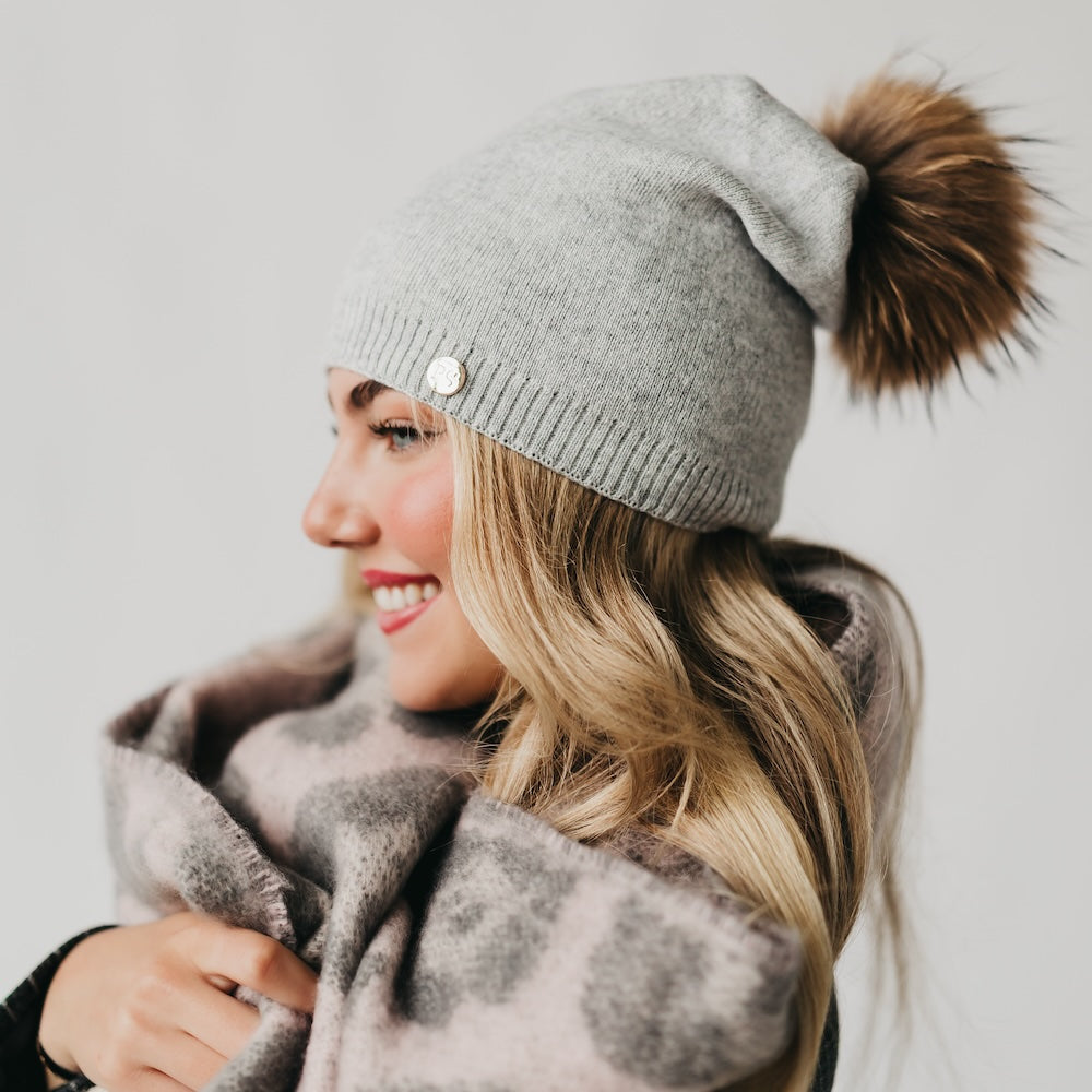 Light gray knit slouchy beanie with ribbed cuff, gold-tone button, and brown fur pom