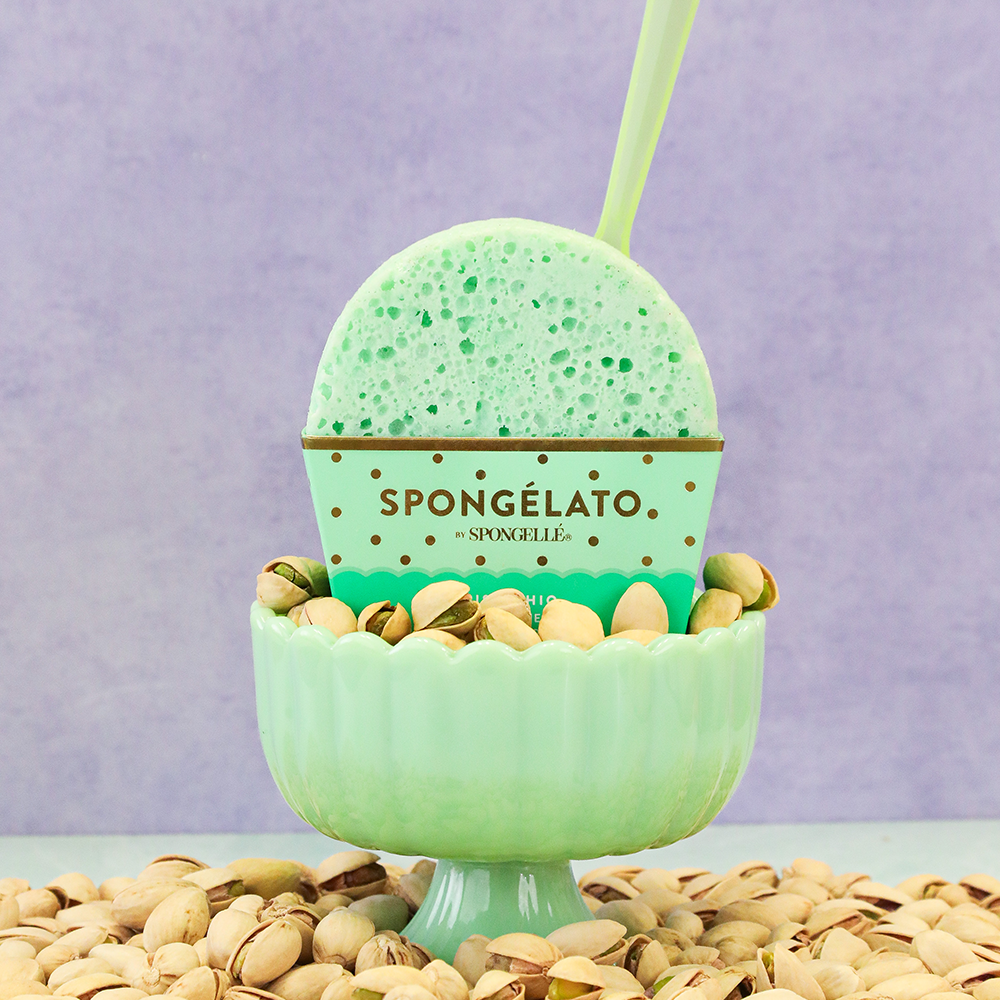 Green textured bath buffer sponge on pedestal, surrounded by pistachio nuts