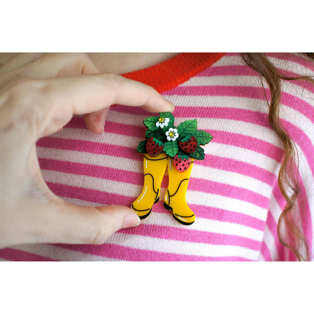 Hand holding: yellow boot strawberry brooch on a pink striped sweater