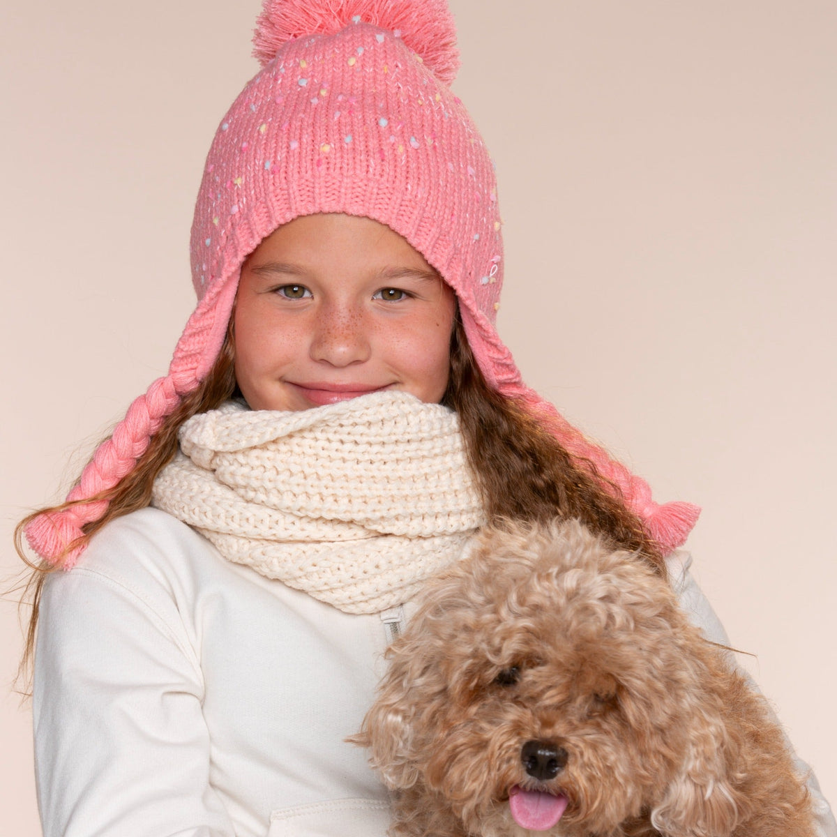 Pink flecked knit hat with earflaps, pom-pom, and braided ties, worn