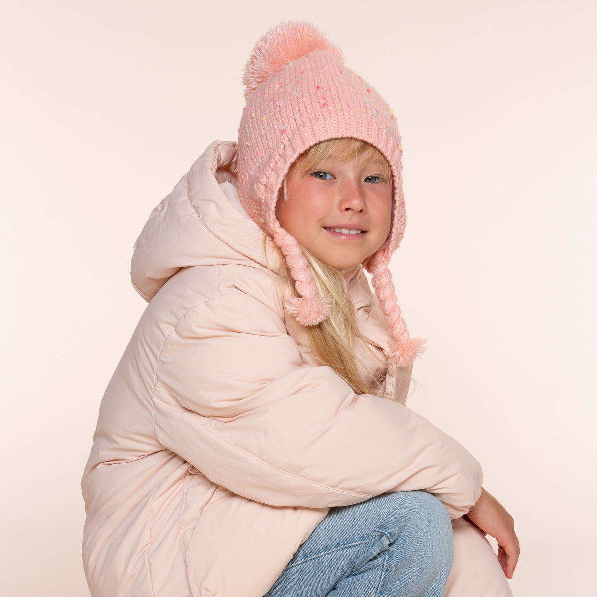 Girls' coral-pink flecked knit earflap hat with pom-pom and braided ties, worn on a child