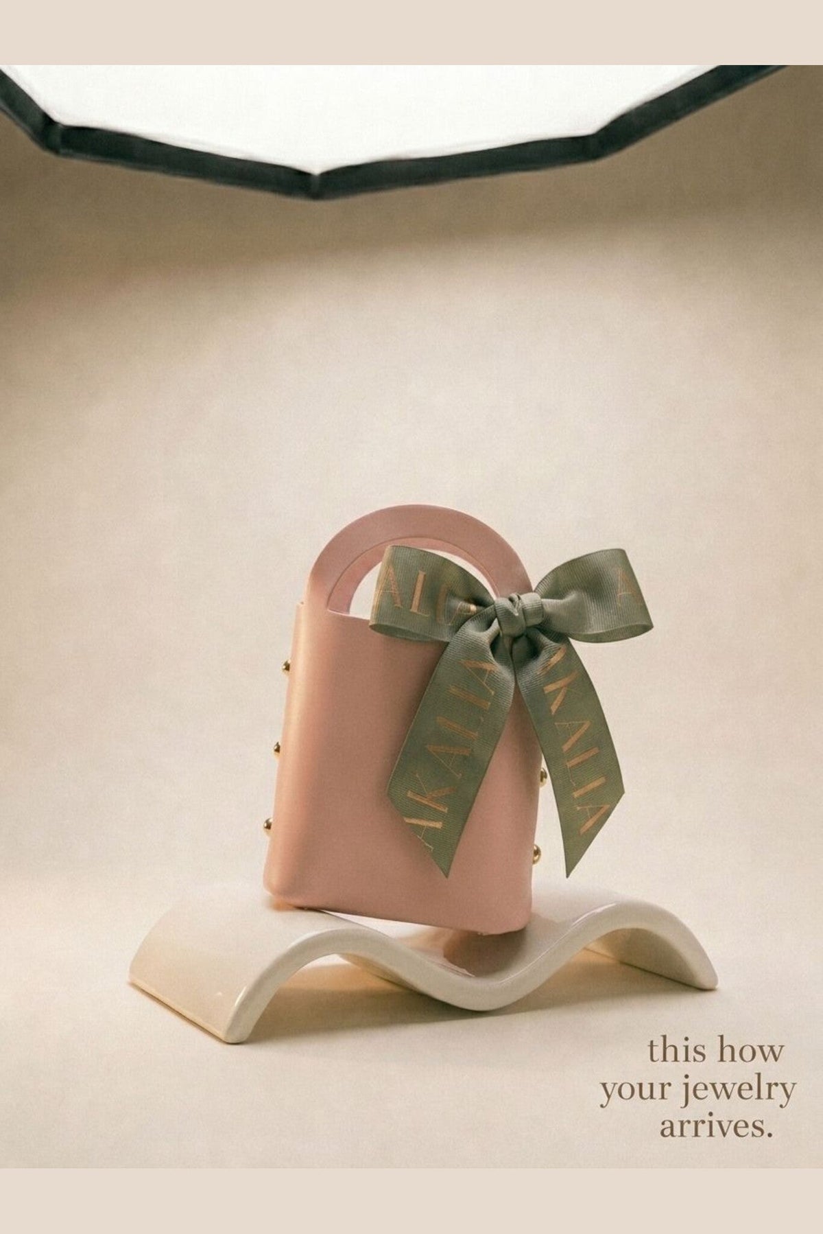 Pink jewelry pouch with green ribbon and AKAKLA logo, shown on white stand