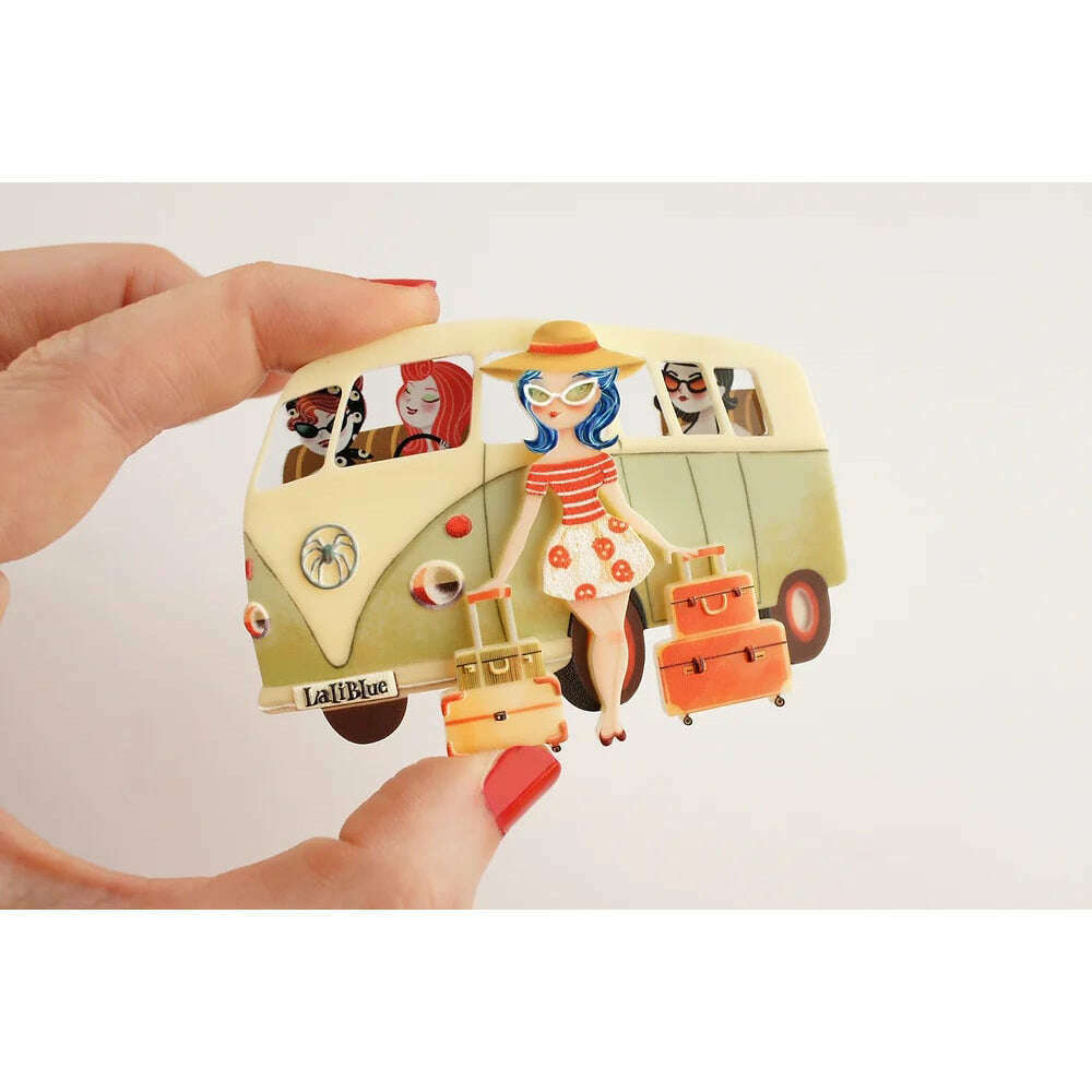 Vintage van brooch held in hand with cream van, blue-haired woman, orange suitcases