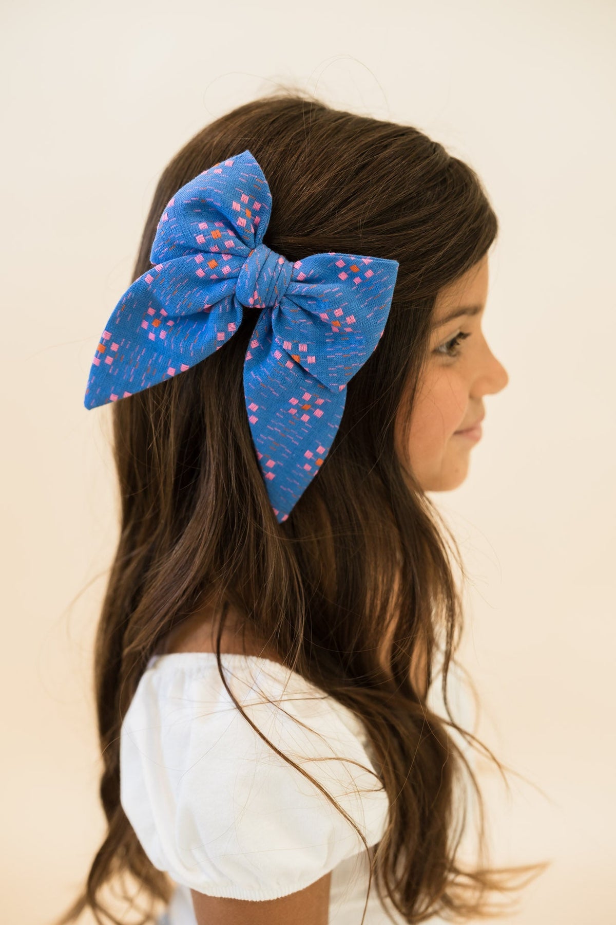 Blue fabric hair bow with multicolor square print, worn on long brown hair.